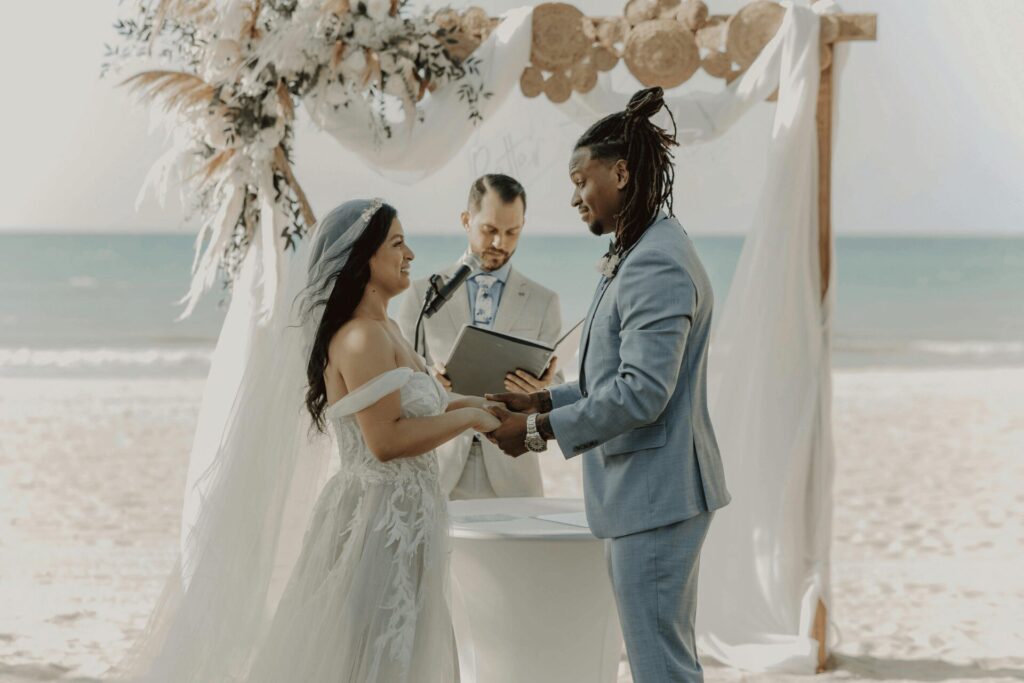 A groom in a light blue suit exchanging vows with his bride at a beach ceremony