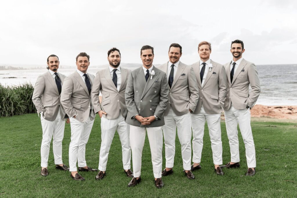 A groom and six groomsmen in matching stone jackets and cream trousers on a lawn by the sea