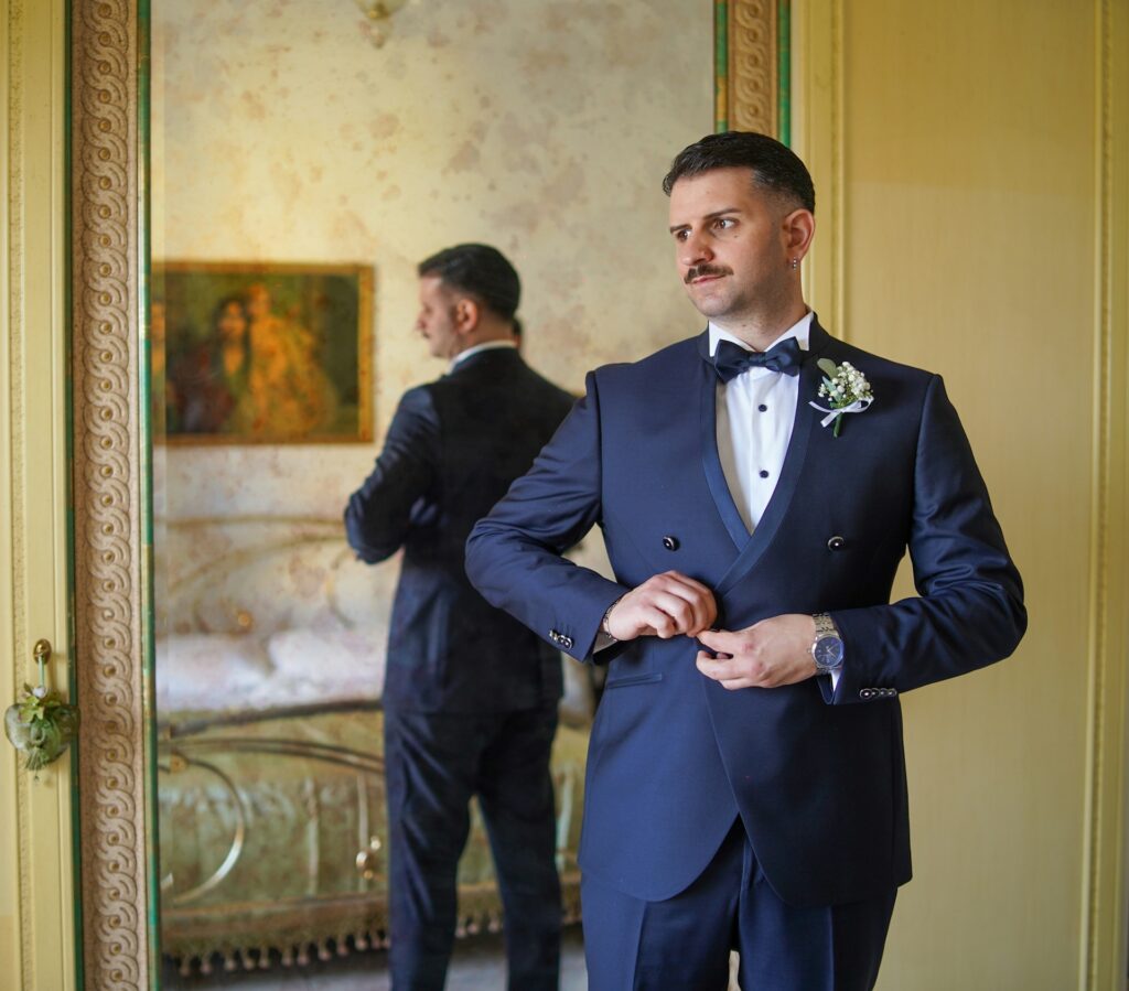 A groom in a navy double-breasted dinner suit, bow tie and buttonhole flower, fastening his jacket in front of a mirror